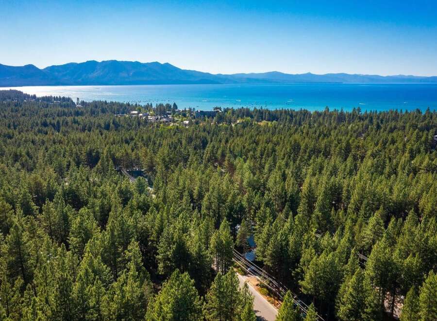 Experience the ultimate lakeside living with this stunning aerial view of a property nestled amidst a dense pine forest, offering breathtaking vistas of a crystal-clear lake and majestic mountains.