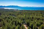 Experience the ultimate lakeside living with this stunning aerial view of a property nestled amidst a dense pine forest, offering breathtaking vistas of a crystal-clear lake and majestic mountains.