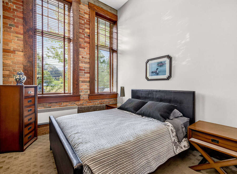 Experience industrial-chic living in this stunning bedroom featuring exposed brick walls and large wooden-framed windows that flood the space with natural light.