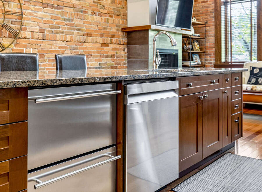 This kitchen island features sleek stainless steel appliances, including a dishwasher and a drawer-style refrigerator, complemented by rich dark wood cabinetry.