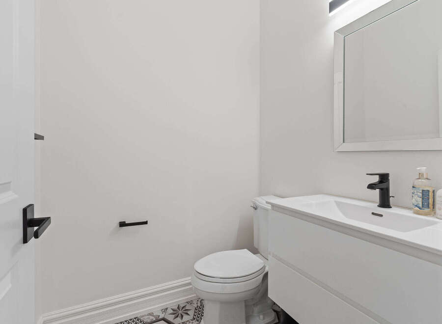 This modern bathroom features a sleek white vanity with a minimalist black faucet, complemented by a matching toilet and a decorative tiled floor.