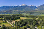 Discover serene living with this stunning aerial view of a picturesque landscape, featuring a tranquil lake, lush green forests, and majestic snow-capped mountains.