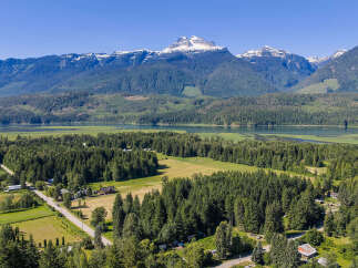 Discover serene living with this stunning aerial view of a picturesque landscape, featuring a tranquil lake, lush green forests, and majestic snow-capped mountains.
