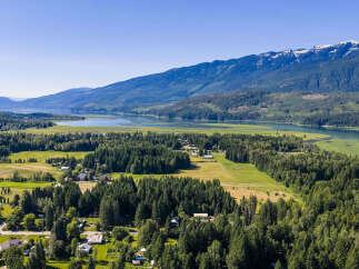 Discover serene living in this picturesque landscape, where lush green fields meet a tranquil lake, all framed by majestic snow-capped mountains.