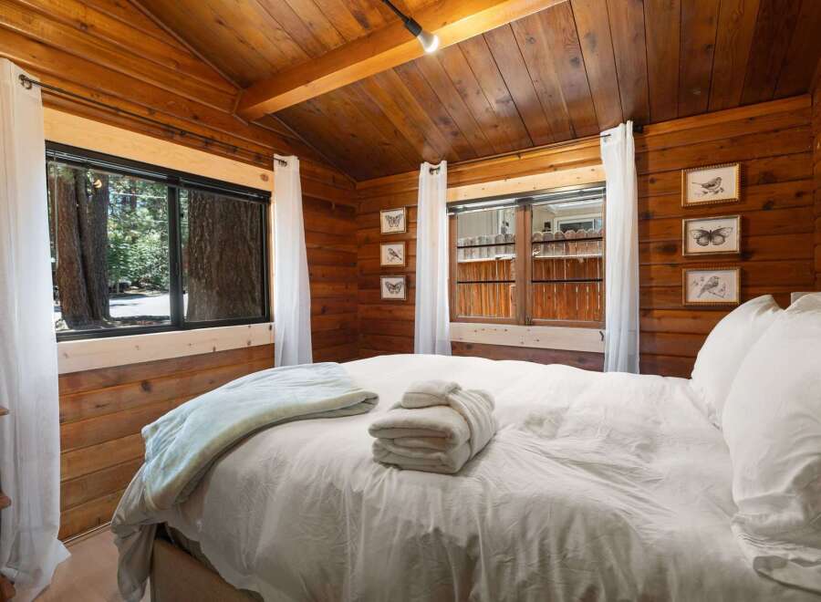Experience rustic charm in this cozy cabin bedroom, featuring warm wood paneling and a comfortable bed adorned with plush white linens and a light blue throw.