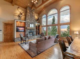 Experience breathtaking mountain views from this stunning living room, featuring a grand stone fireplace and soaring vaulted ceilings with exposed wooden beams.