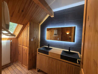 Experience rustic charm in this bathroom featuring a wooden ceiling and walls, complemented by a sleek vanity with double black sinks and a modern illuminated mirror.