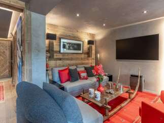 Experience luxury mountain living in this stunning chalet living room, featuring a cozy grey sofa adorned with vibrant red and charcoal pillows, set against a rustic wood-paneled wall.
