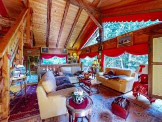 Experience rustic charm in this cozy wooden cabin living room, featuring plush sofas adorned with faux fur throws, a traditional fireplace, and large windows offering serene forest views.