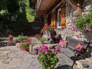 Charming stone cottage with a rustic wooden balcony and shutters, surrounded by vibrant flower gardens.