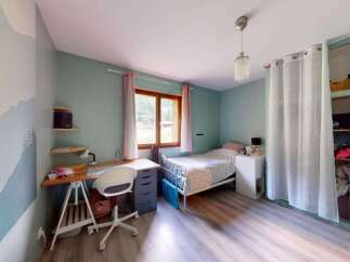 This charming bedroom features a cozy single bed with a patterned duvet, a functional wooden desk with a white swivel chair, and a large window offering natural light.