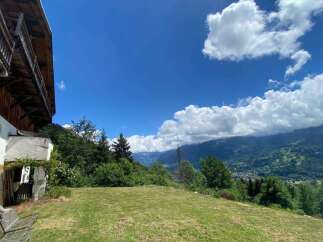 Experience breathtaking mountain views from this charming property, featuring a rustic wooden balcony and a lush green lawn perfect for outdoor relaxation.