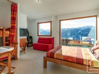 Experience breathtaking mountain views from this charming studio apartment, featuring a cozy bunk bed area, a vibrant red sofa, and a modern wood-burning stove.