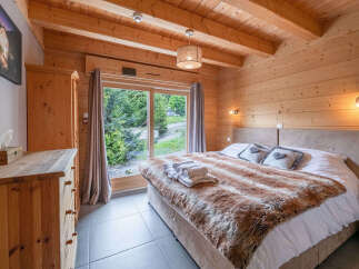 Experience cozy mountain living in this charming wooden bedroom, featuring a plush fur throw and a large window with stunning forest views.