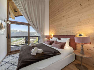 Experience breathtaking mountain views from this luxurious bedroom, featuring a large window with a balcony overlooking a charming village.