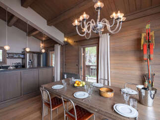 Experience rustic charm in this dining area, featuring a large wooden table set for a meal, complemented by a modern chandelier and warm wood paneling.