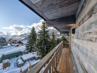 Experience the charm of a winter wonderland from this rustic balcony, offering breathtaking views of snow-covered mountains and a quaint village below.