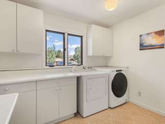 This laundry room features ample white cabinetry for storage, a large countertop space, and a window with a scenic view of the outdoors.