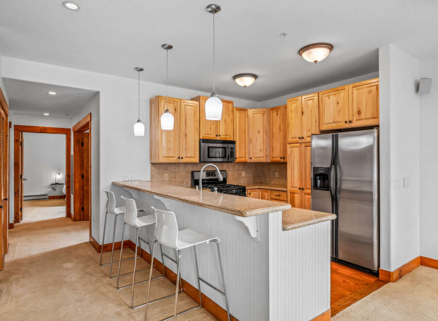 This spacious kitchen features light wood cabinetry, a granite countertop breakfast bar with white stools, and stainless steel appliances including a refrigerator and microwave.