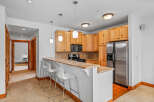 This spacious kitchen features light wood cabinetry, a granite countertop breakfast bar with white stools, and stainless steel appliances including a refrigerator and microwave.