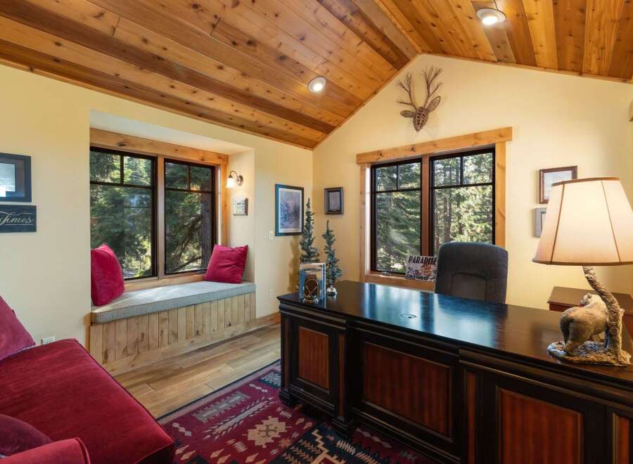 This rustic home office features a large wooden desk with a decorative lamp, complemented by a cozy window seat overlooking a serene forest view.