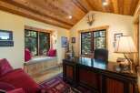 This rustic home office features a large wooden desk with a decorative lamp, complemented by a cozy window seat overlooking a serene forest view.