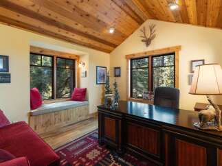 This rustic home office features a large wooden desk with a decorative lamp, complemented by a cozy window seat overlooking a serene forest view.