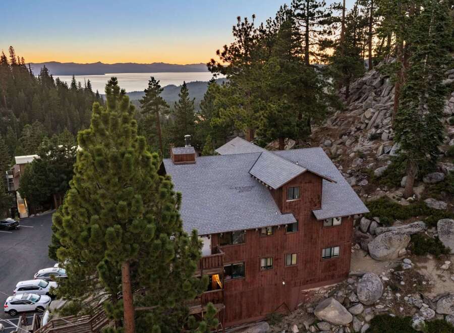 Nestled amidst towering pine trees and rocky terrain, this rustic wooden lodge offers breathtaking views of a serene lake and distant mountains at sunset.
