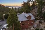 Nestled amidst towering pine trees and rocky terrain, this rustic wooden lodge offers breathtaking views of a serene lake and distant mountains at sunset.