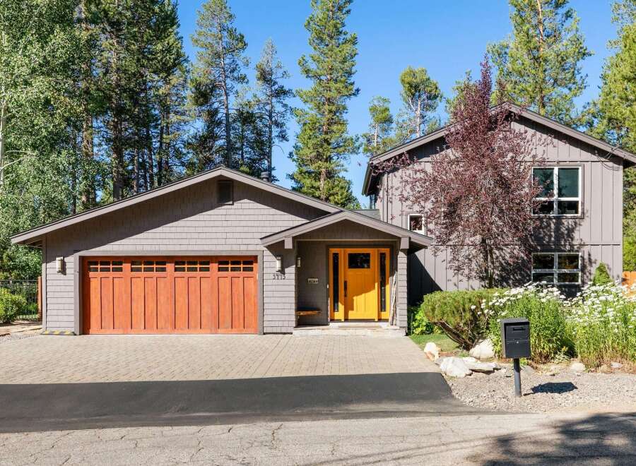 Discover this charming mountain home featuring a striking orange front door and a warm wooden garage door, set against a backdrop of tall pine trees.