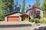 Discover this charming mountain home featuring a striking orange front door and a warm wooden garage door, set against a backdrop of tall pine trees.