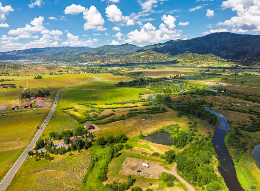 Experience the serene beauty of this expansive rural landscape, featuring a winding river, lush green fields, and majestic mountains in the distance.
