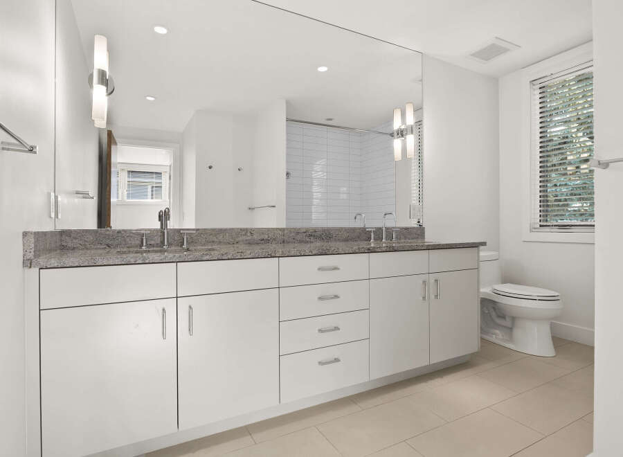 This modern bathroom features a sleek white vanity with a granite countertop and dual sinks, complemented by a large mirror reflecting a tiled shower.
