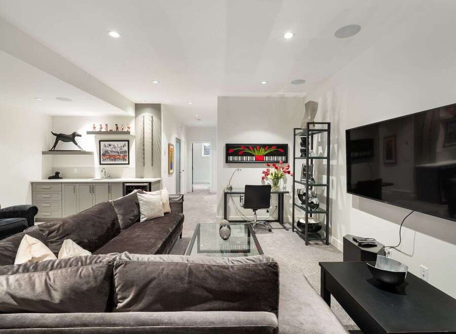 This modern basement living area features a spacious sectional sofa, a sleek wet bar with ample cabinetry, and a dedicated workspace with a glass-top desk.