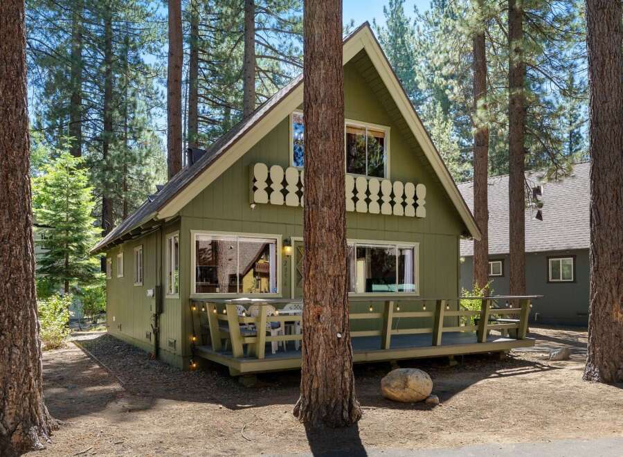 Charming green cabin nestled among tall pine trees, featuring a welcoming front porch with outdoor seating and string lights.