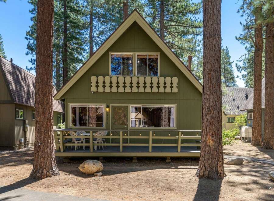Charming green cabin nestled among tall pine trees, featuring a welcoming front porch with outdoor seating and string lights.