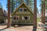 Charming green cabin nestled among tall pine trees, featuring a welcoming front porch with outdoor seating and string lights.