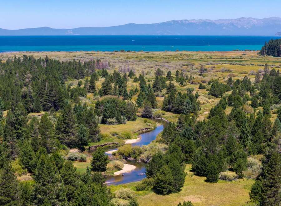 Discover this stunning property featuring a serene river winding through lush green trees and meadows, leading to a breathtaking view of a vast blue lake and distant mountains.