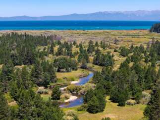 Discover this stunning property featuring a serene river winding through lush green trees and meadows, leading to a breathtaking view of a vast blue lake and distant mountains.