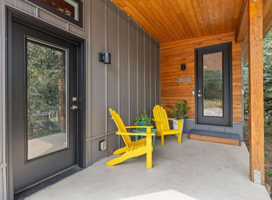 Welcome guests to this modern home with a charming covered porch featuring two bright yellow Adirondack chairs and lush potted plants.