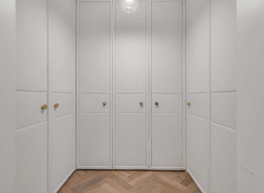 Step into this elegant walk-in closet featuring custom built-in cabinetry with textured white panels and sleek brass hardware.