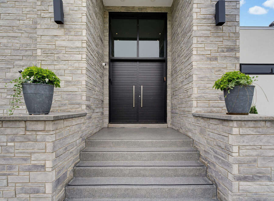 This modern home features a striking entrance with a double black door and a large transom window, framed by textured grey stone walls.