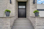 This modern home features a striking entrance with a double black door and a large transom window, framed by textured grey stone walls.