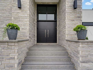 This modern home features a striking entrance with a double black door and a large transom window, framed by textured grey stone walls.