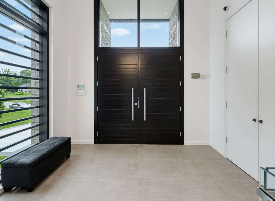 Step into a grand foyer featuring striking black double doors, complemented by a large window with horizontal slats offering a glimpse of the lush outdoors.