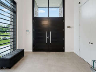 Step into a grand foyer featuring striking black double doors, complemented by a large window with horizontal slats offering a glimpse of the lush outdoors.