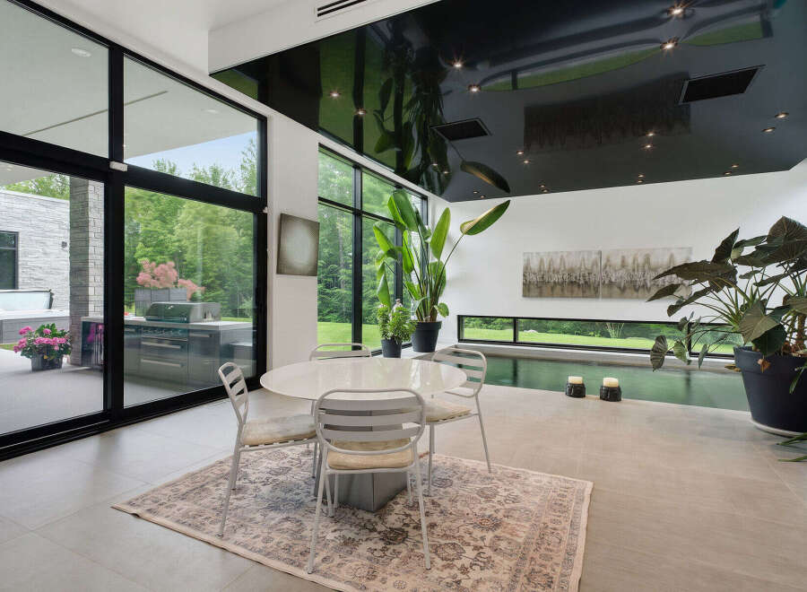 Experience modern luxury in this stunning indoor-outdoor living space, featuring a sleek dining area with a glass-top table and chairs, complemented by a sophisticated outdoor kitchen and hot tub visible through expansive glass doors.