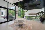 Experience modern luxury in this stunning indoor-outdoor living space, featuring a sleek dining area with a glass-top table and chairs, complemented by a sophisticated outdoor kitchen and hot tub visible through expansive glass doors.