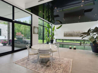 Experience modern luxury in this stunning indoor-outdoor living space, featuring a sleek dining area with a glass-top table and chairs, complemented by a sophisticated outdoor kitchen and hot tub visible through expansive glass doors.