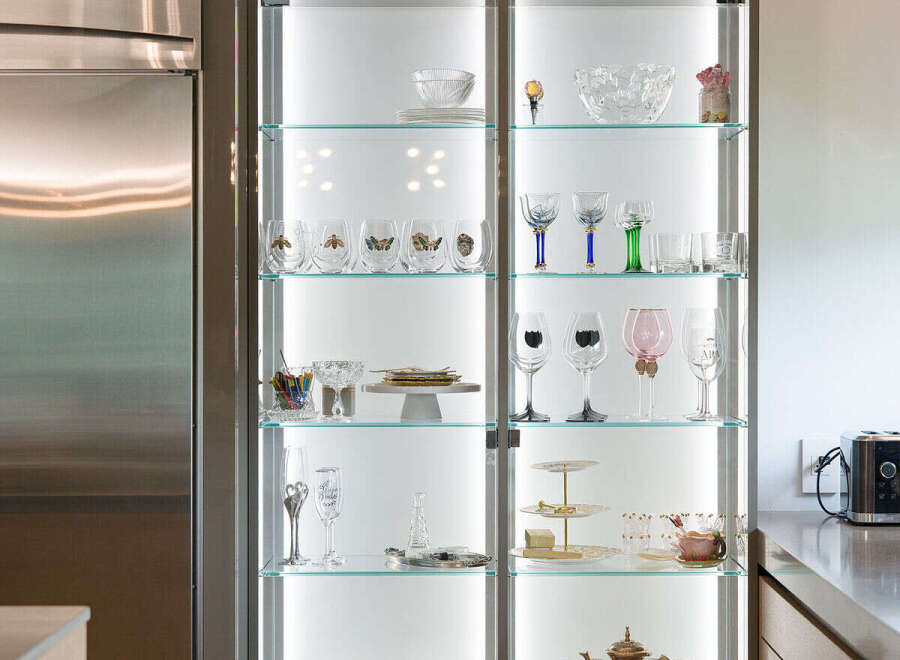 Showcasing a modern kitchen with a sleek, illuminated glass display cabinet filled with elegant glassware and decorative items.